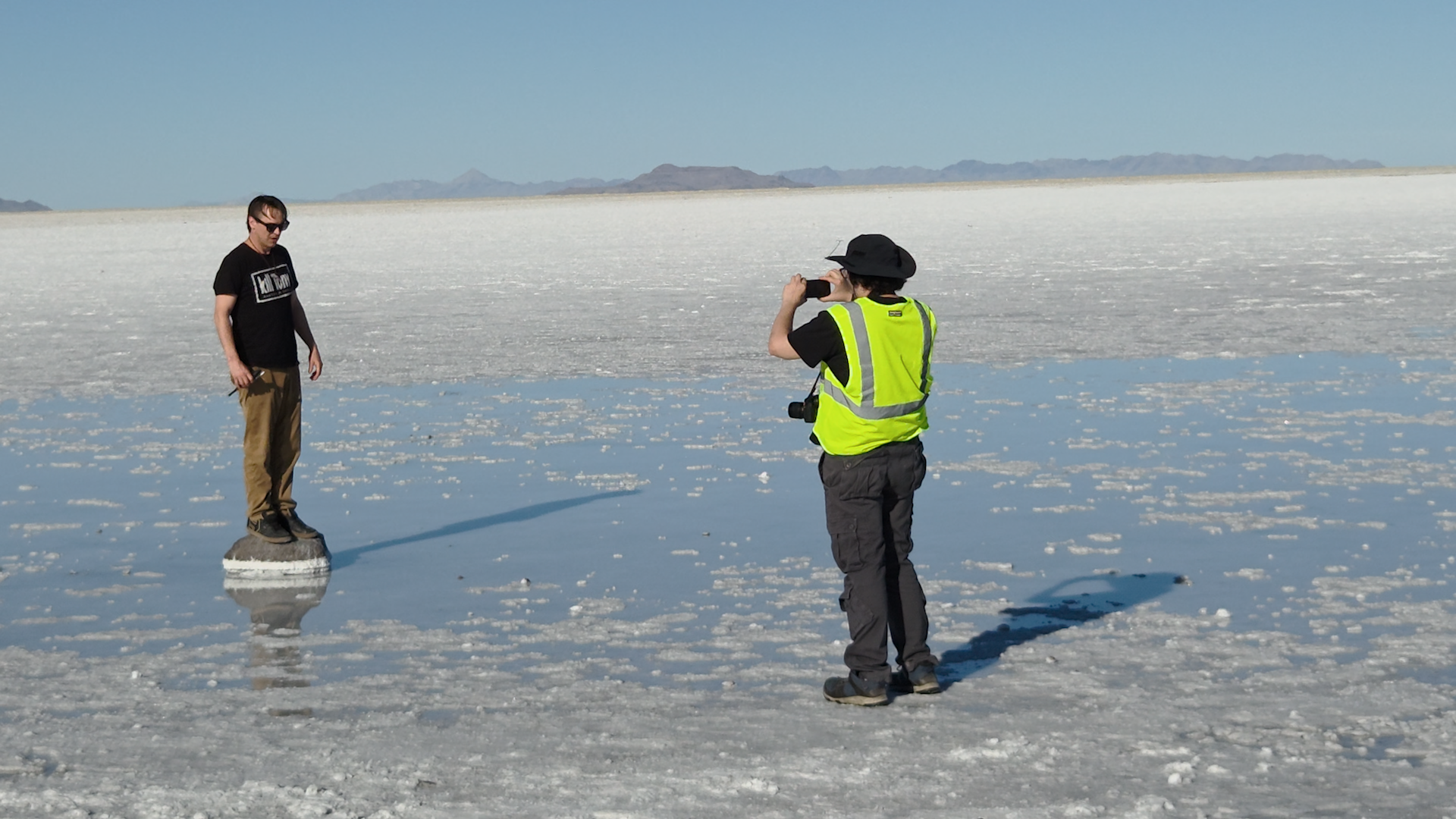 A man standing on a rock while a man in a yellow safety vest sets up a shot at an outdoor stop in the Bonneville salt flats.